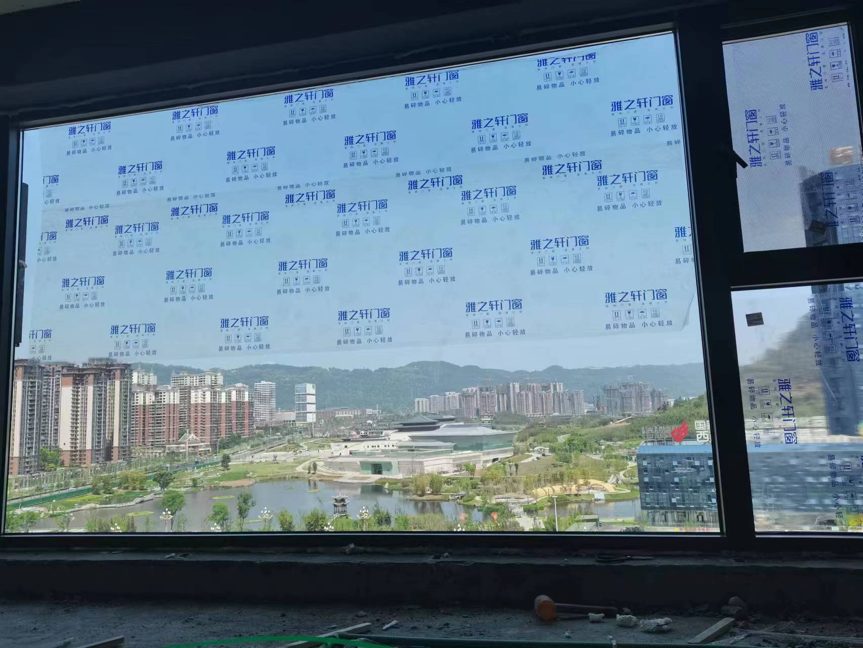 室内装修施工现场,黑色边框的雅之轩品牌大窗户已安装到位。玻璃表面贴满印有“雅之轩门窗”及“易碎物品 小心轻放”字样的蓝色保护膜,有效防止施工刮花。窗外视野极佳,