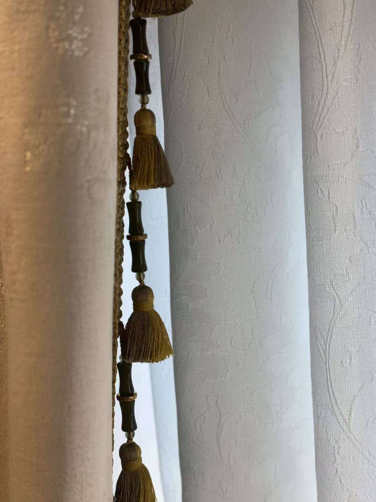 这是一张展示精致窗帘细节的特写图片。画面主体为垂坠感极佳的窗帘流苏边饰，采用金棕色丝线编织成流苏穗子，上方串联深墨绿色的竹节状装饰珠，极具复古韵味。左侧主帘布料