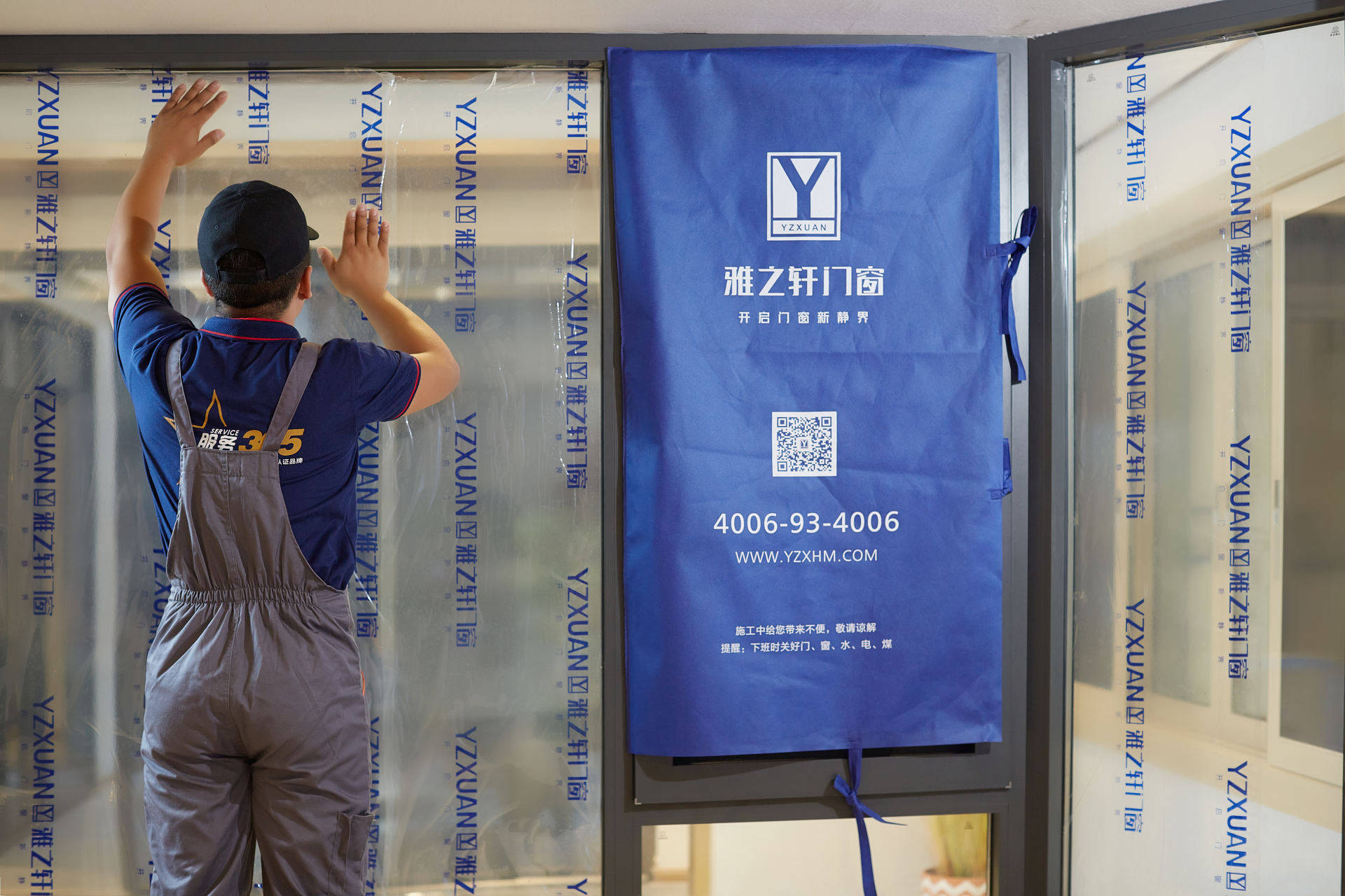 雅之轩门窗施工现场实拍，身穿蓝色工装的安装师傅正在仔细粘贴带有品牌Logo的窗户保护膜。深灰色窗框搭配大面积玻璃，中间悬挂蓝色施工遮挡布，印有品牌标识、服务热线