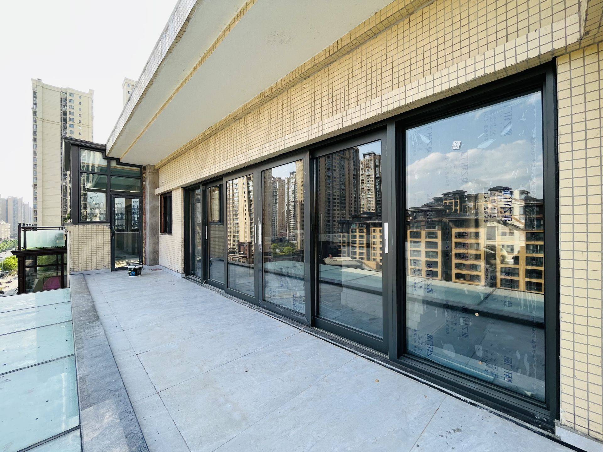 这张图片展示了一个高层住宅的阳台封闭工程实拍。主体为深灰色断桥铝材质的落地推拉门，安装在米黄色外墙砖背景下，大面积玻璃设计保证了极佳的采光与视野。玻璃表面覆盖着